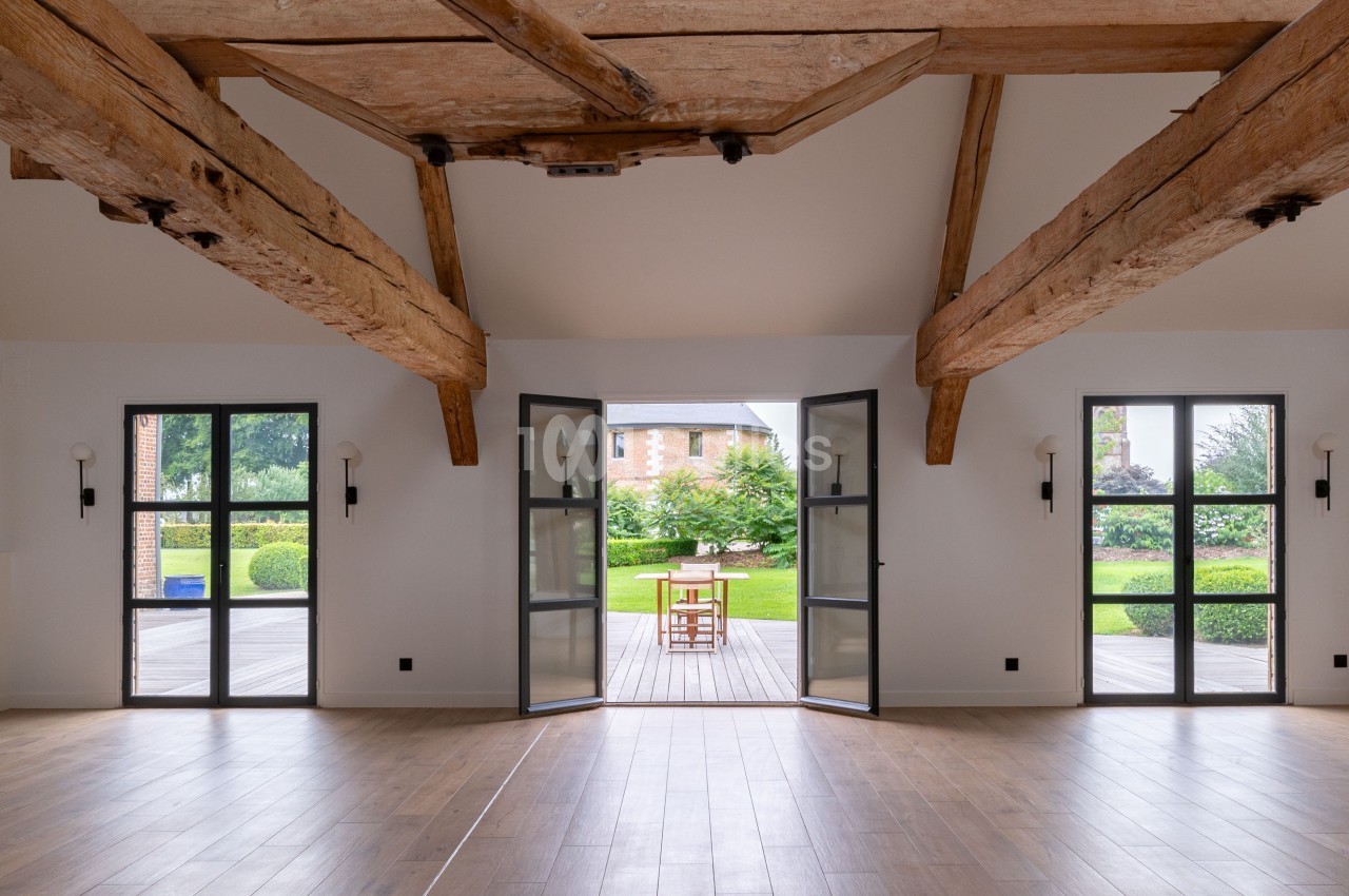 Vue intérieure d'une pièce lumineuse avec poutres apparentes, parquet, portes vitrées ouvertes sur un jardin.