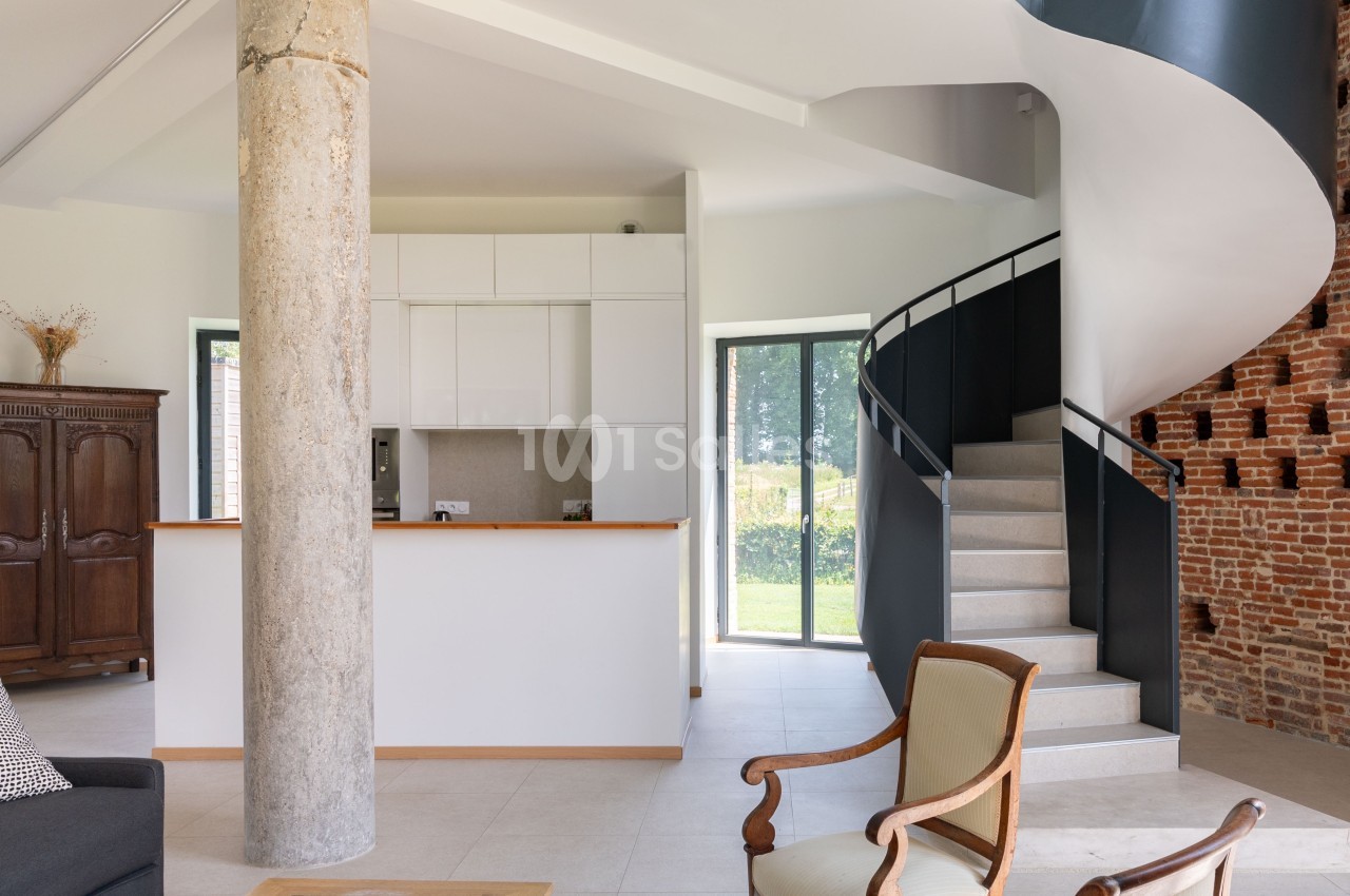 Intérieur lumineux avec escalier en colimaçon, colonne en béton, meubles classiques et vue sur un jardin.