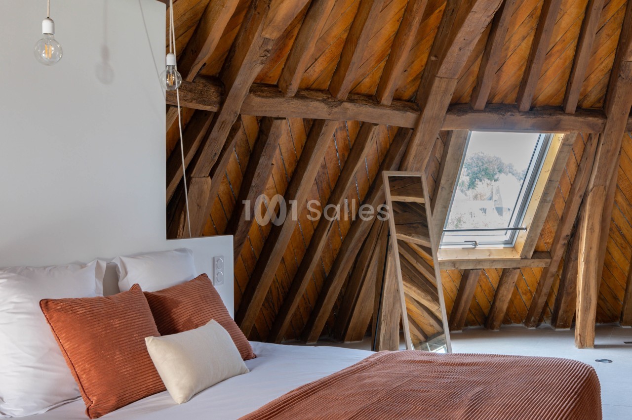 Chambre sous combles avec lit double, couverture orange, poutres apparentes et fenêtre de toit laissant entrer la lumière.