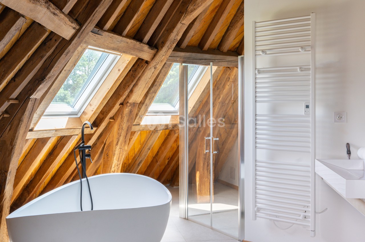 Salle de bain sous combles avec baignoire îlot blanche, poutres apparentes, douche vitrée et sèche-serviettes mural.