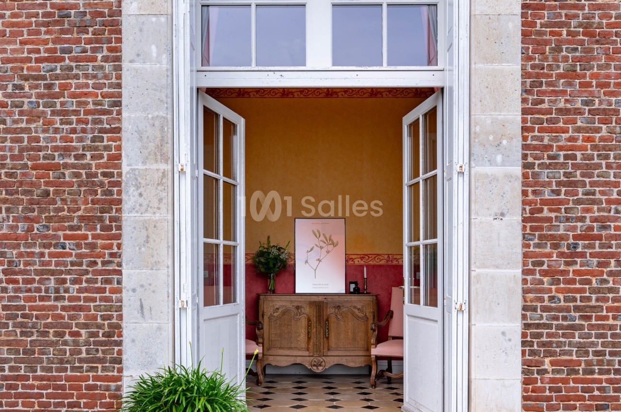 Porte-fenêtre ouverte sur une pièce intérieure avec un meuble ancien, une plante et un sol en damier.