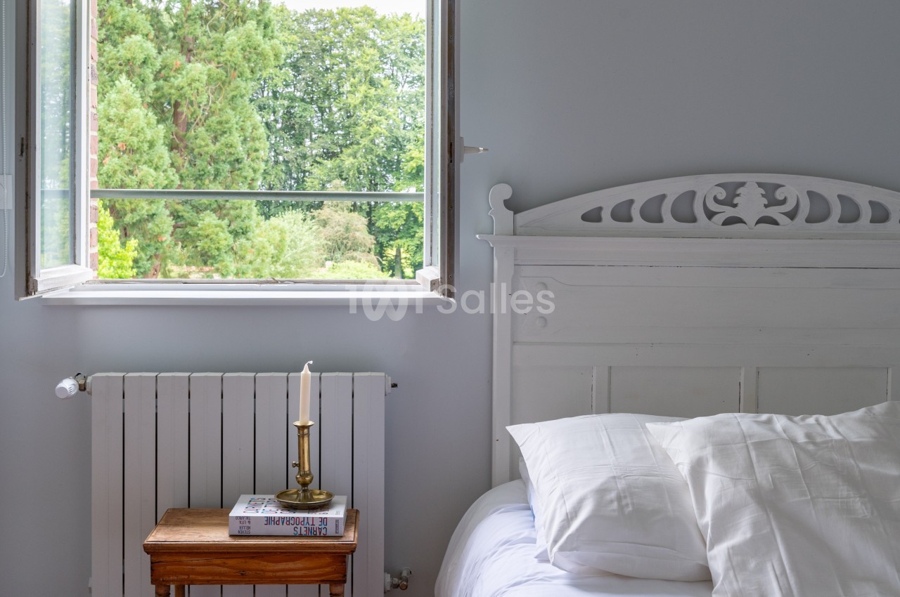 Chambre lumineuse avec un lit blanc, une table de chevet en bois, une bougie et une fenêtre ouverte sur un jardin verdoyant.