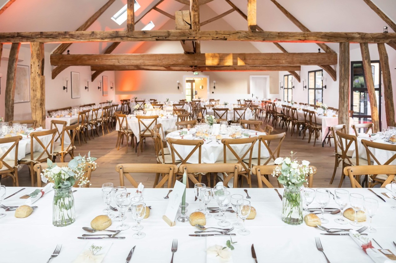 Salle de réception lumineuse avec tables dressées, décorée de fleurs et poutres apparentes en bois.