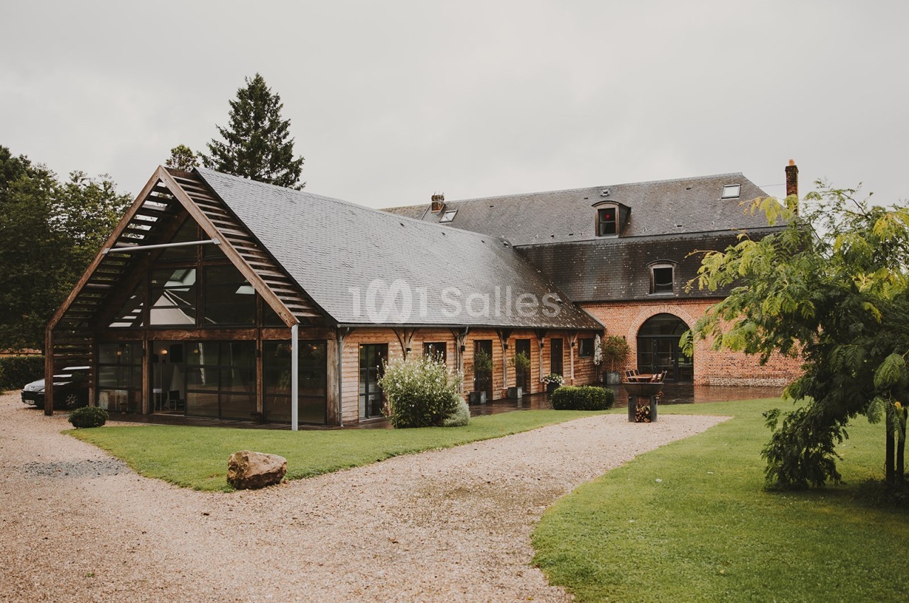 Bâtiment en briques et bois avec toit en ardoise, entouré de pelouse et d'arbres, sous un ciel nuageux.