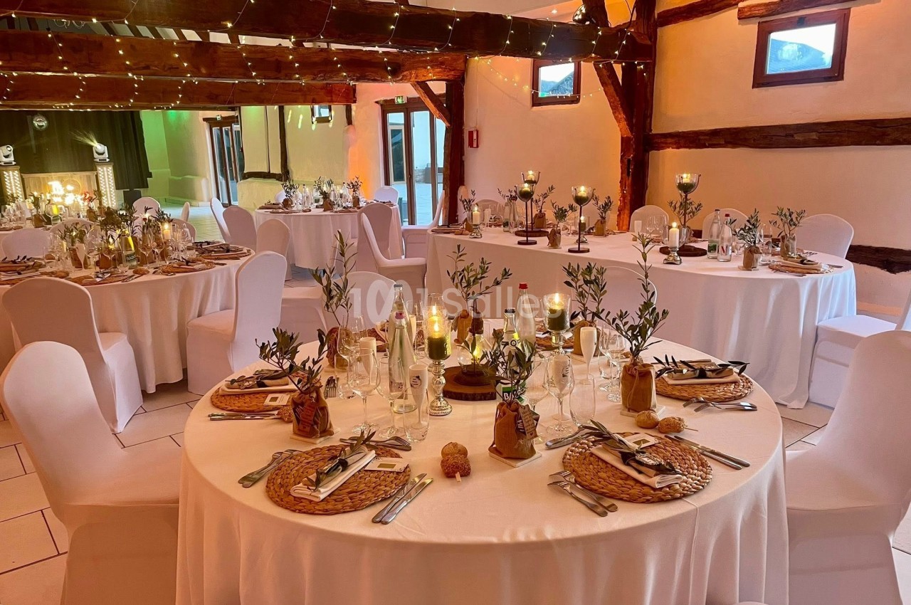 Salle de réception décorée avec des tables rondes, nappes blanches, bougies et éléments végétaux dans une ambiance…