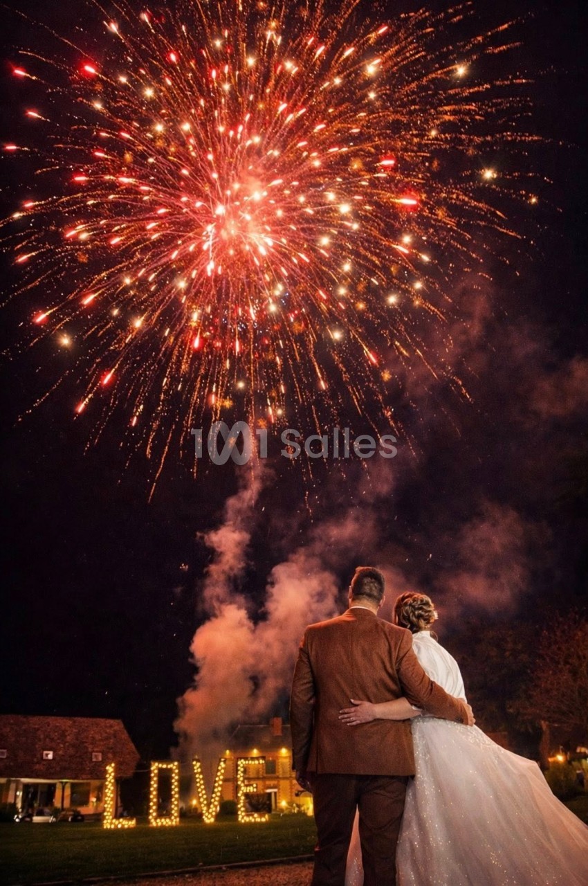 Un couple observe un feu d'artifice éclatant dans le ciel nocturne, avec le mot ’LOVE’ illuminé en arrière-plan.