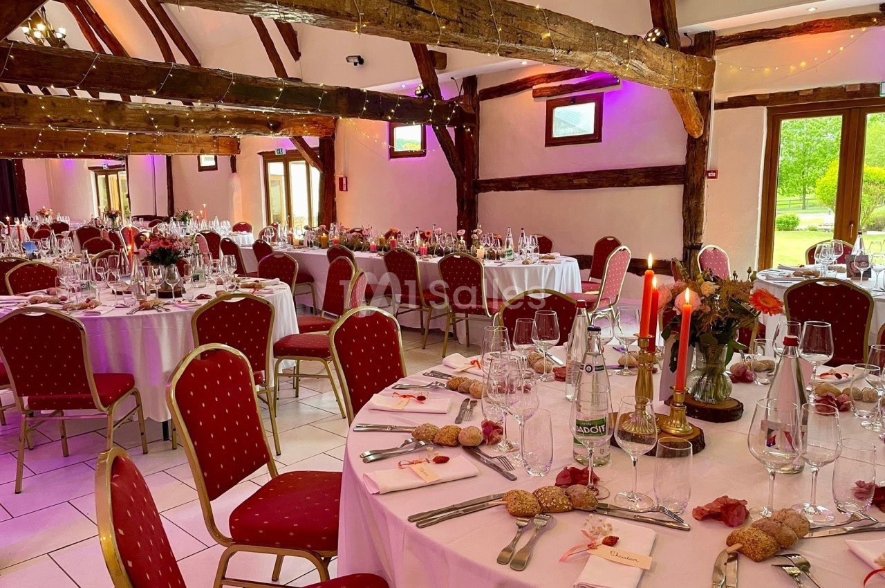 Salle de réception décorée avec des tables rondes dressées, chaises rouges et lumières suspendues.