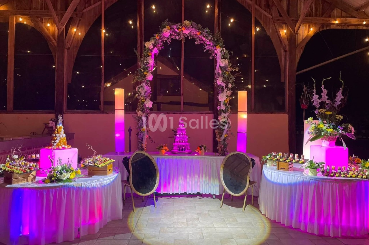 Salle décorée pour un événement avec arche florale, tables garnies et éclairage rose et chaleureux.