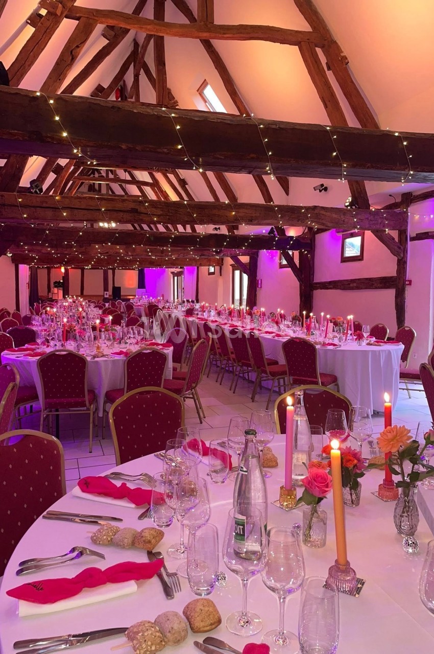 Salle de réception décorée avec des tables dressées, bougies allumées et éclairage d'ambiance rose sous une charpente en…