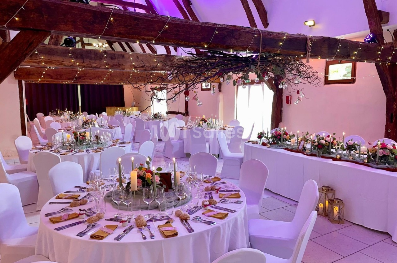Salle de réception décorée pour un mariage avec tables rondes et rectangulaires, bougies et guirlandes lumineuses.