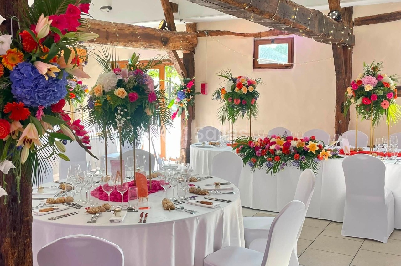 Salle de réception décorée avec des tables rondes, nappes blanches et compositions florales colorées.