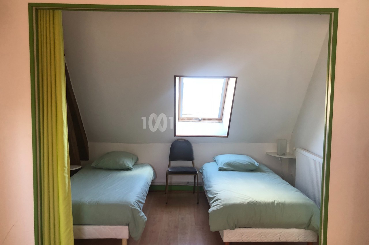 Deux lits simples avec draps verts dans une chambre mansardée, éclairée par une fenêtre de toit.