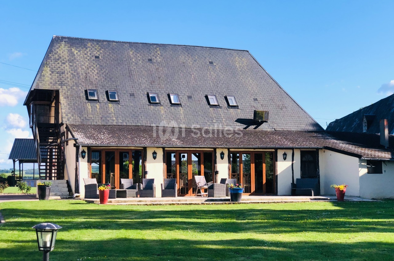 Grande maison avec toit en ardoise, nombreuses fenêtres et terrasse aménagée, entourée d'une pelouse verte.
