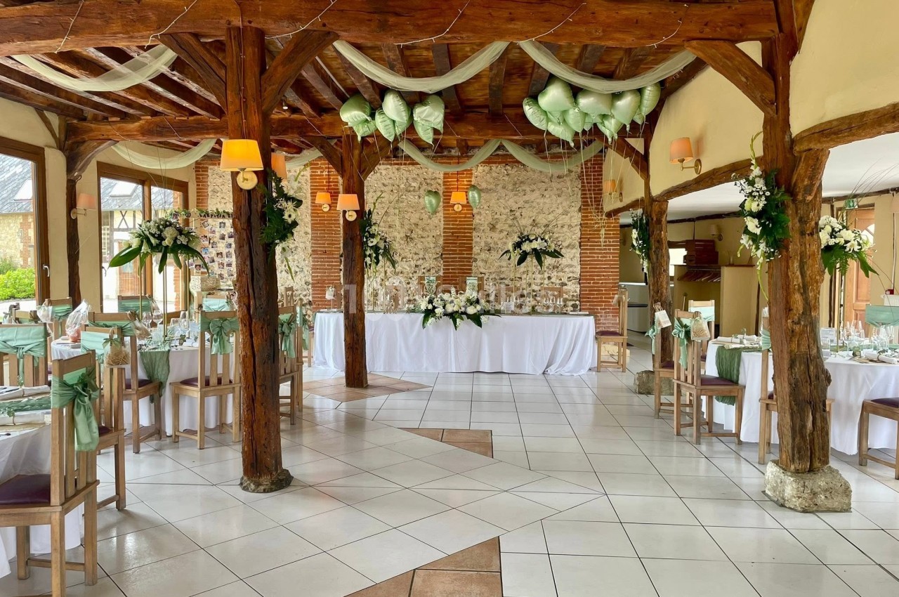 Salle de réception décorée pour un mariage, avec tables dressées, fleurs blanches et vertes, et poutres en bois apparentes.