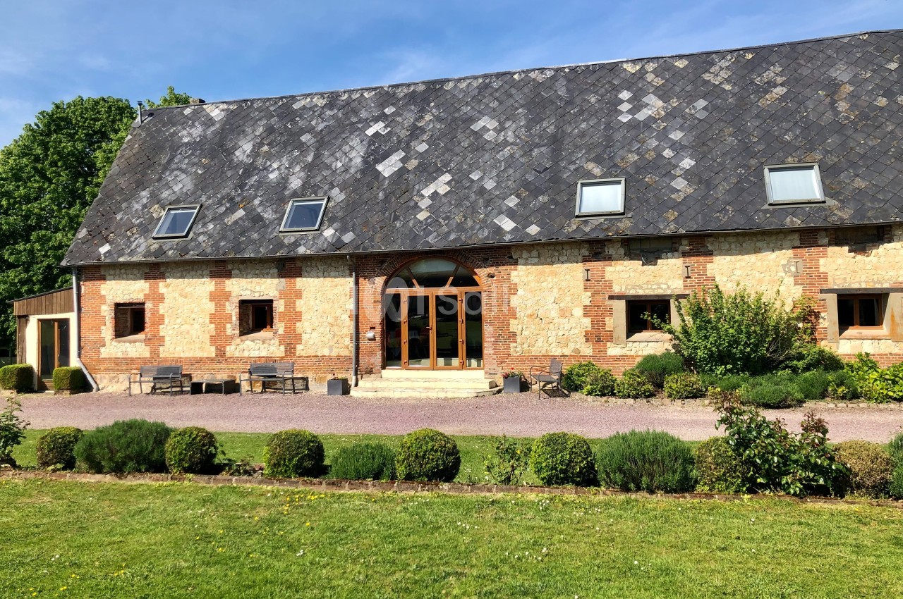 Façade d'une grande maison en briques et pierres avec toit en ardoise, entourée de pelouse et d'arbustes.