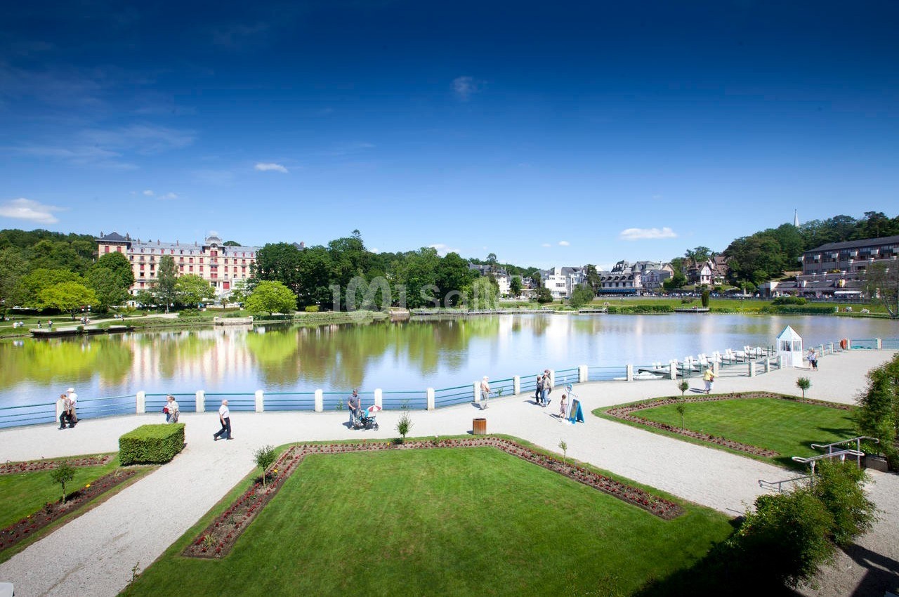 Vue d'un parc avec pelouses, allées, lac entouré de barrières blanches et bâtiments en arrière-plan sous un ciel bleu.
