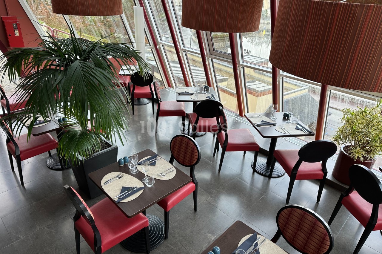 Salle de restaurant lumineuse avec grandes baies vitrées, tables dressées et chaises rouges, plantes en décoration.
