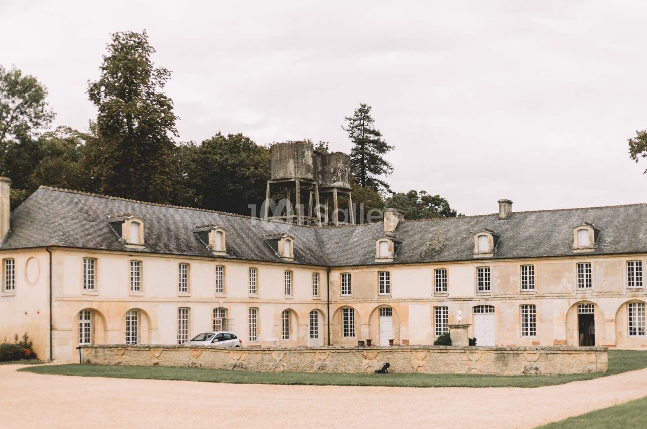 Bâtiment ancien en pierre avec toit en ardoise, cour centrale gravillonnée et structure en bois en arrière-plan.