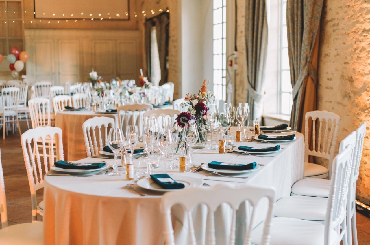 Salle de réception décorée avec des tables rondes dressées, nappes blanches, chaises blanches et arrangements floraux.