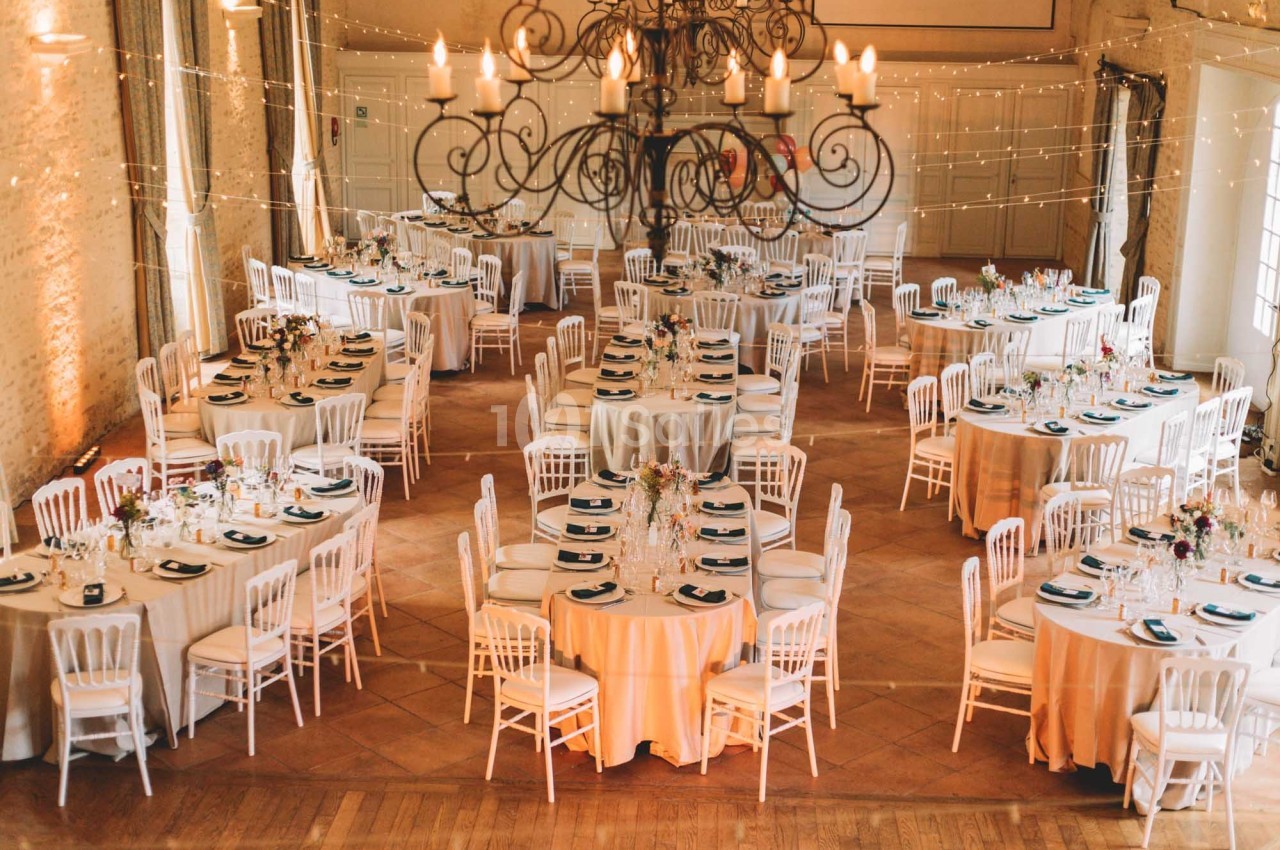 Salle de réception décorée avec des tables rondes dressées, des chaises blanches et un grand lustre central.