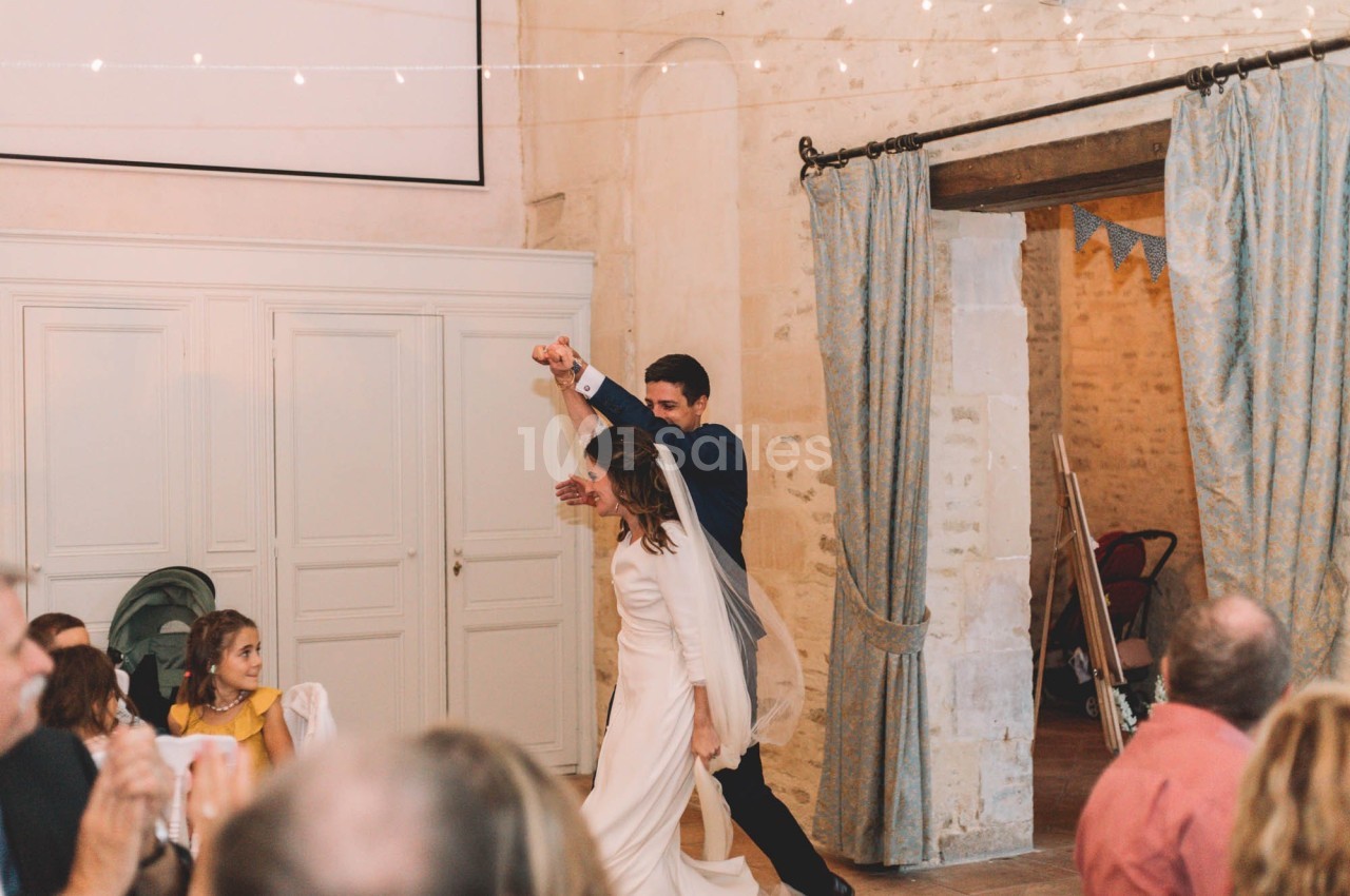 Un couple de mariés danse dans une salle décorée, sous des guirlandes lumineuses, entouré d'invités assis.