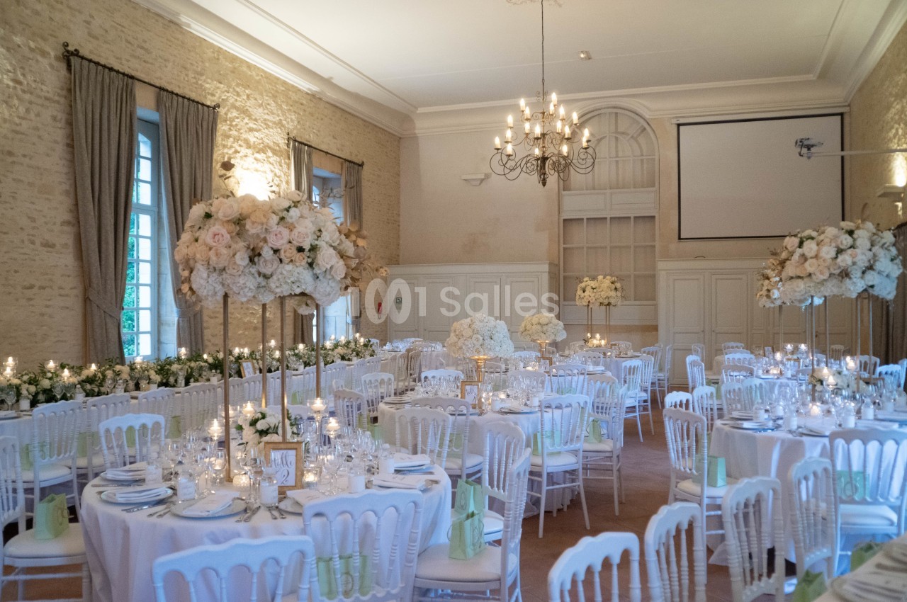 Salle de réception élégante décorée pour un mariage, avec tables dressées, chandeliers et arrangements floraux blancs.