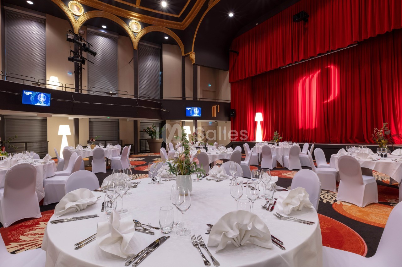 Salle de réception élégante avec tables rondes dressées, nappes blanches et rideaux rouges en arrière-plan.