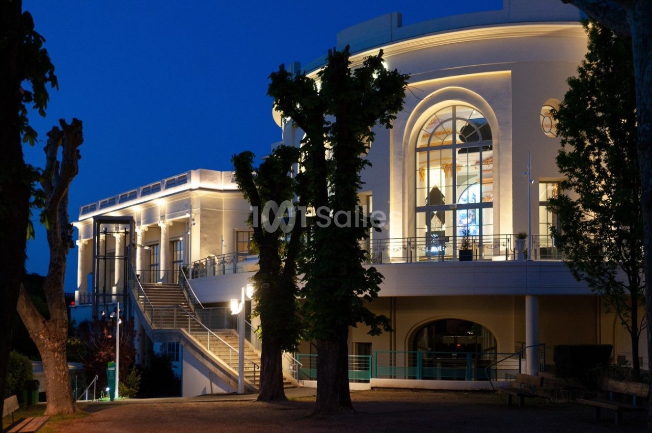 Façade illuminée d'un bâtiment élégant avec de grandes fenêtres et des arbres au premier plan, photographiée de nuit.