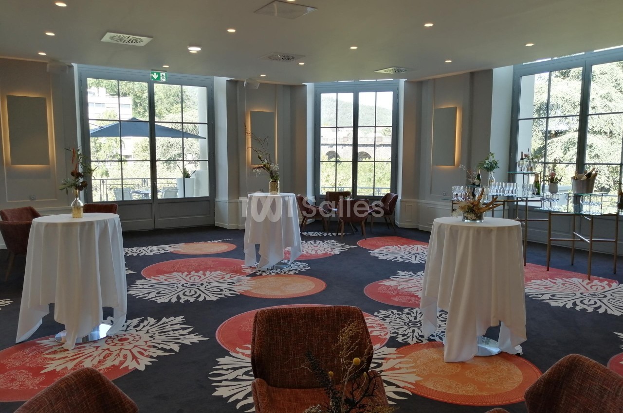 Salle lumineuse avec tables hautes drapées de blanc, chaises, décor floral et grandes fenêtres donnant sur un paysage…