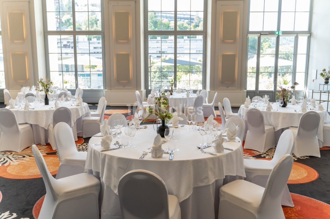 Salle lumineuse avec tables rondes dressées pour un événement, nappes blanches et chaises couvertes.