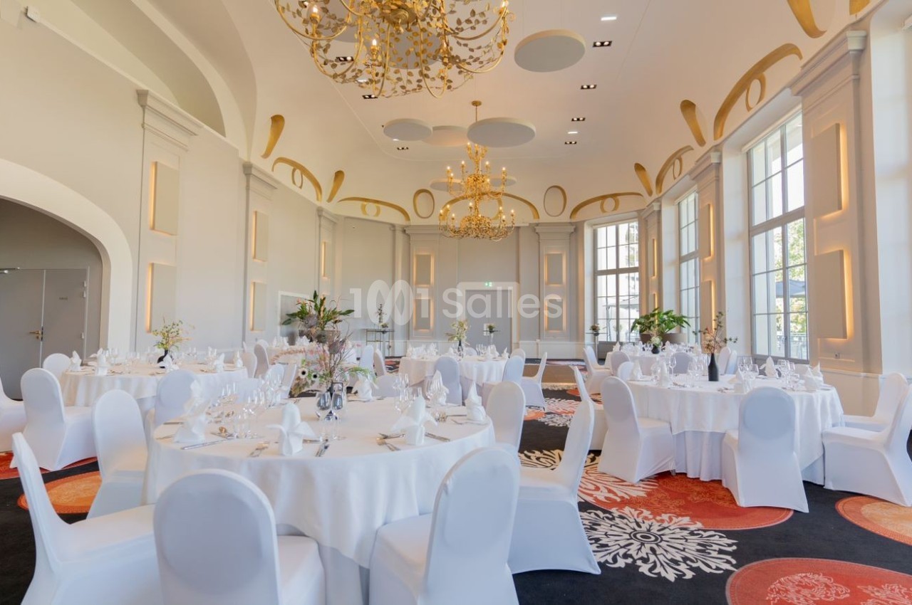 Salle de réception élégante avec tables rondes dressées, chaises blanches et lustres dorés sous un plafond voûté.