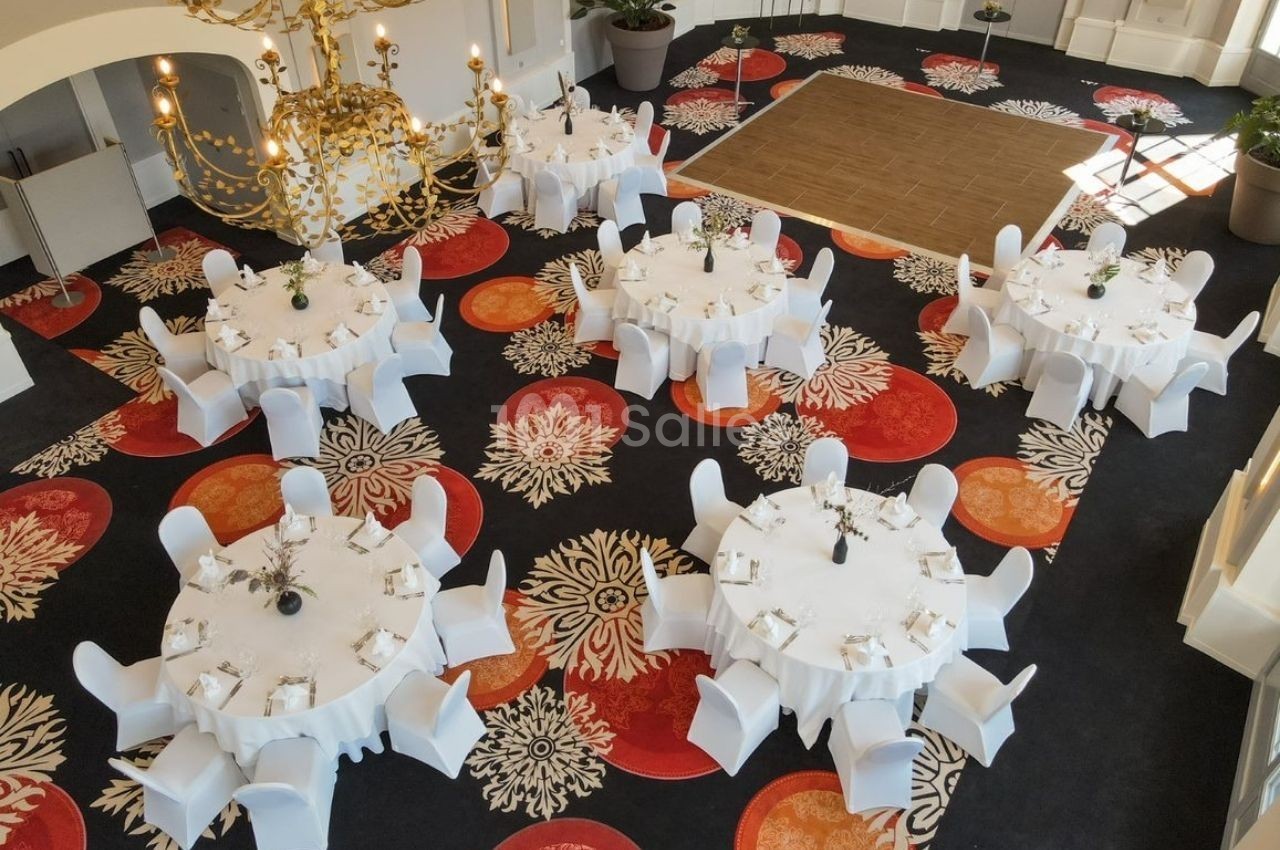 Salle de réception avec tables rondes dressées de nappes blanches, chaises assorties et décor floral au sol.