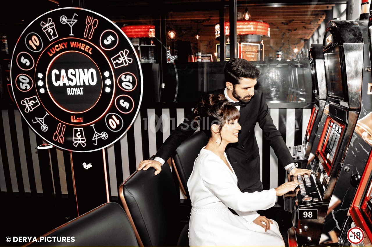 Un homme et une femme jouent à une machine à sous dans un casino, avec une roue de la chance en arrière-plan.