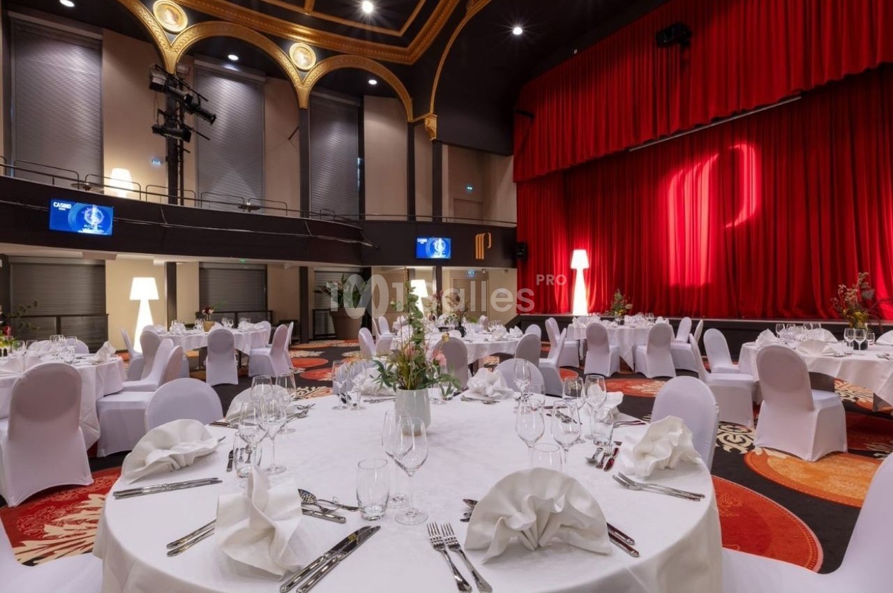 Salle de réception élégante avec tables rondes dressées, chaises blanches et rideaux rouges sur une scène.