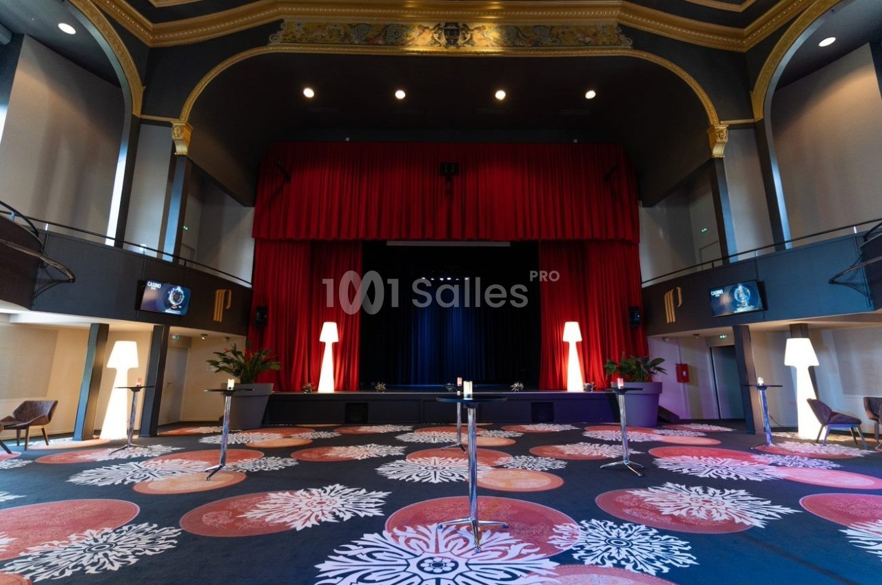 Salle de spectacle avec rideaux rouges, scène centrale, plafond orné et moquette colorée à motifs floraux.