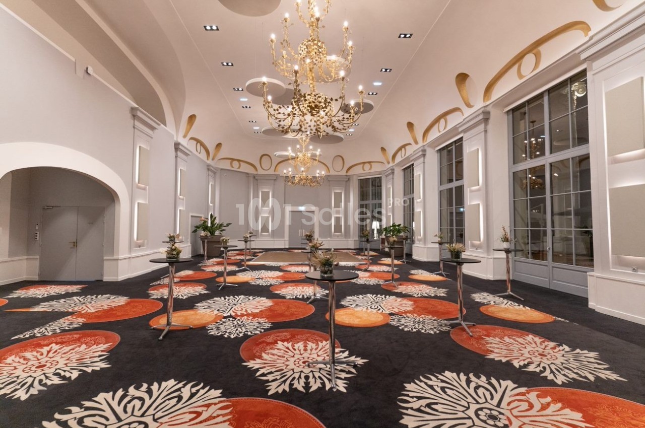 Salle élégante avec lustres dorés, grandes fenêtres, tapis coloré à motifs floraux et tables hautes dispersées.