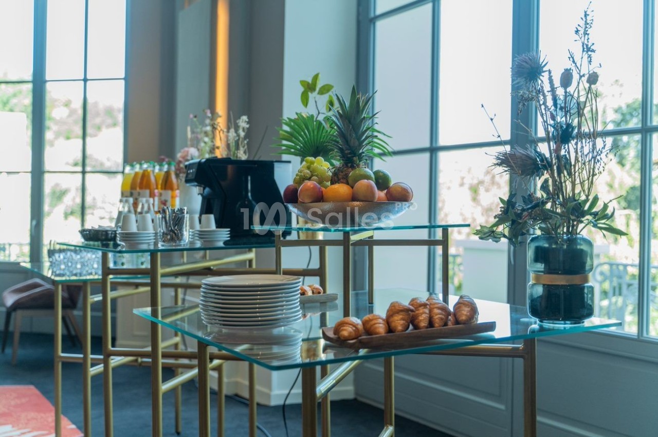 Buffet avec fruits, viennoiseries, boissons et vaisselle sur des tables en verre près de grandes fenêtres lumineuses.