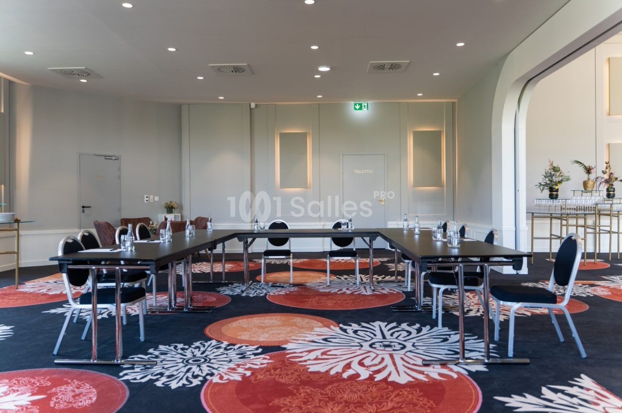 Salle de réunion lumineuse avec tables en U, chaises noires, bouteilles d'eau et décor floral sur tapis coloré.