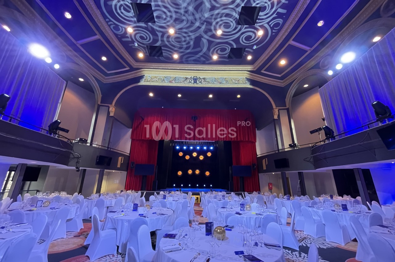 Salle de réception élégante avec tables dressées, éclairage bleu et scène ornée de rideaux rouges.
