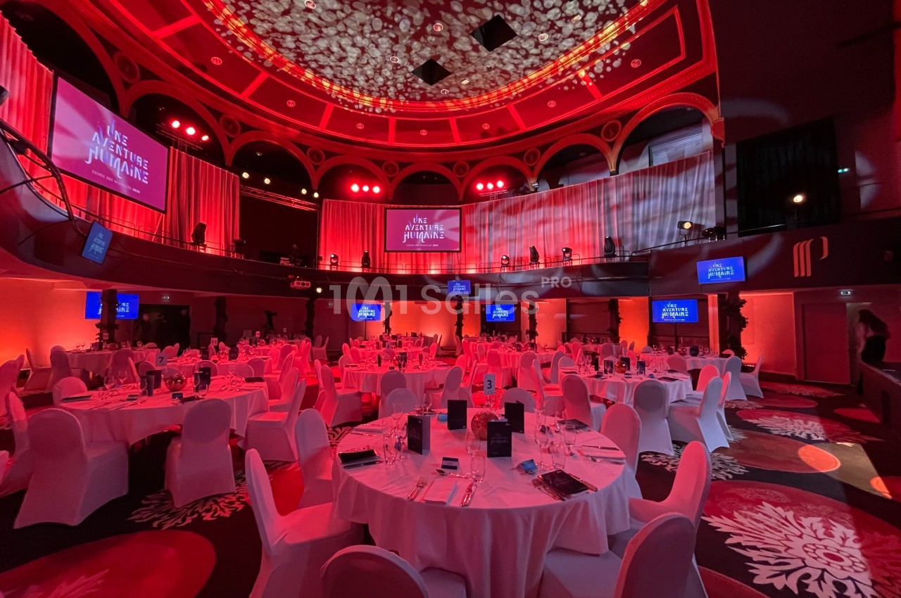 Salle de réception éclairée en rouge avec tables rondes dressées pour un événement, écrans affichant un titre.