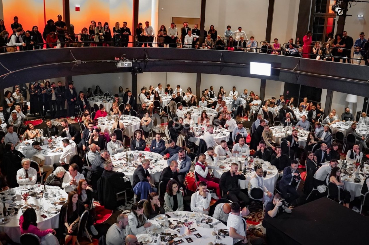 Salle comble avec des invités assis autour de tables rondes lors d'un événement, sous un éclairage chaleureux.