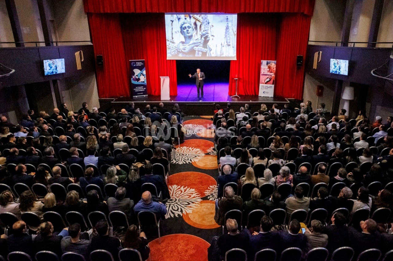 Salle de conférence avec un orateur sur scène devant un public, écran projetant une image artistique en arrière-plan.