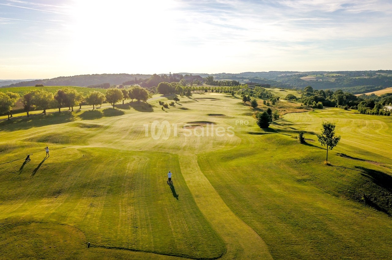 Vue aérienne d'un terrain de golf verdoyant avec des arbres alignés et un paysage vallonné sous un ciel ensoleillé.