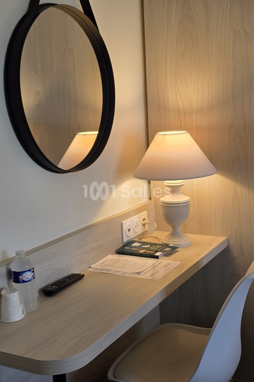 Bureau en bois clair avec une chaise blanche, une lampe allumée, un miroir rond, des documents et une bouteille d'eau.