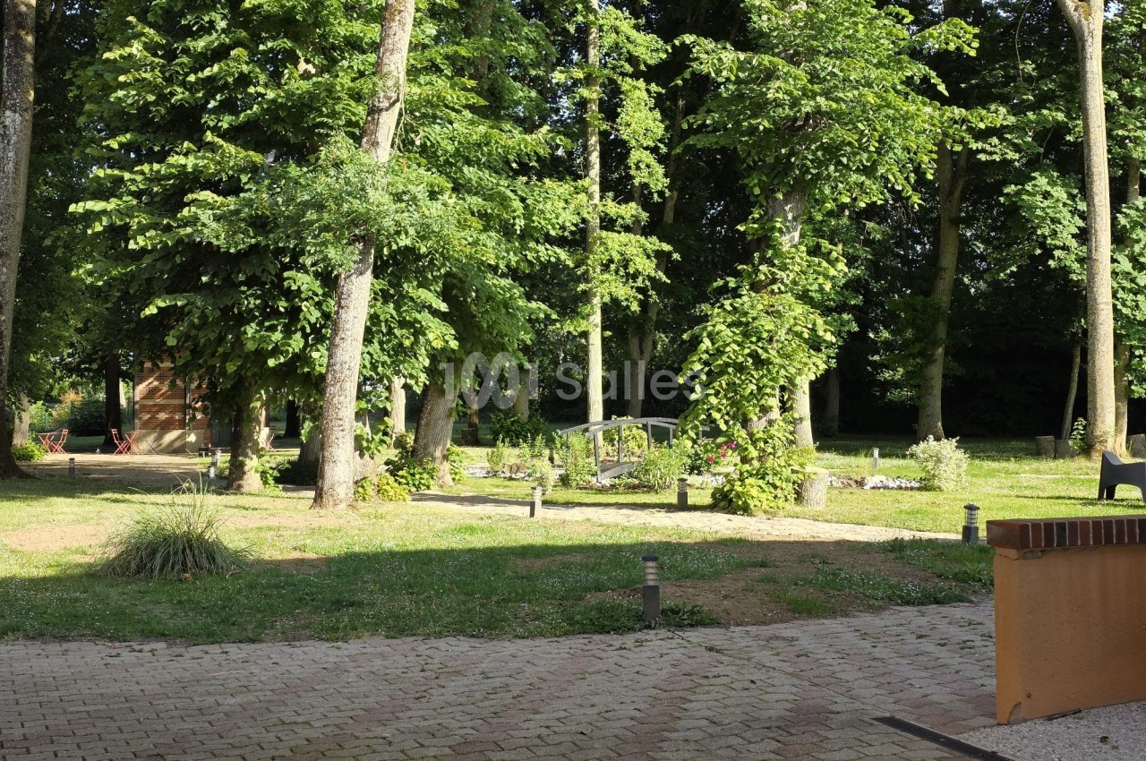 Vue d'un jardin arboré avec des arbres, des plantes et des chemins pavés sous une lumière ensoleillée.