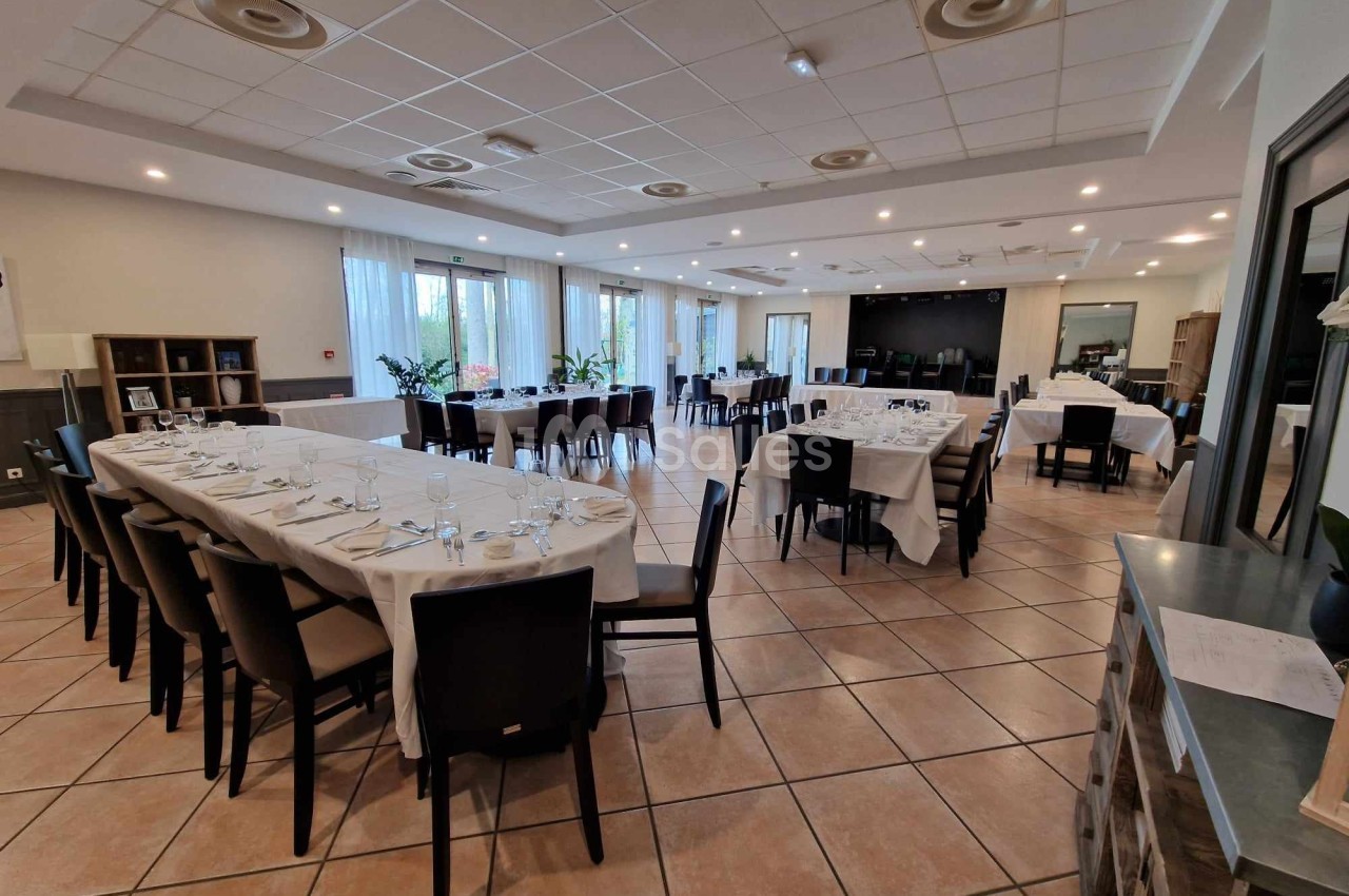 Salle de réception lumineuse avec tables dressées, nappes blanches et chaises noires, prête pour un événement.