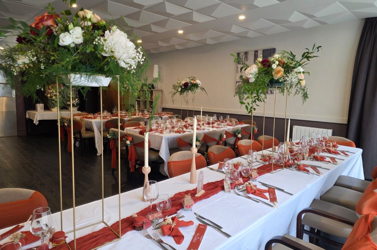 Salle de réception décorée avec des tables dressées, nappes blanches, chemins de table rouges et compositions florales.