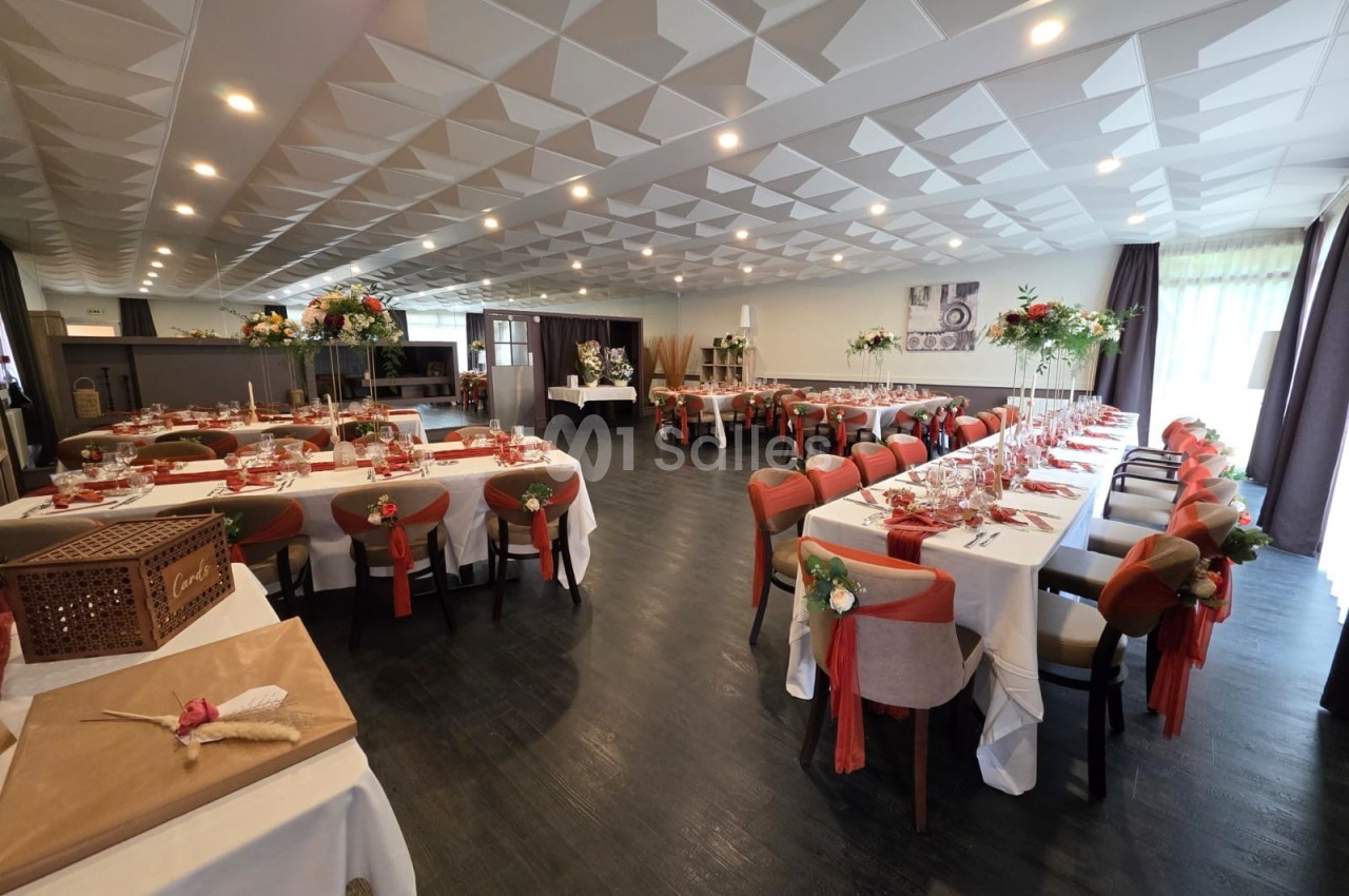 Salle de réception décorée avec des tables dressées, chaises ornées de rubans rouges et arrangements floraux.
