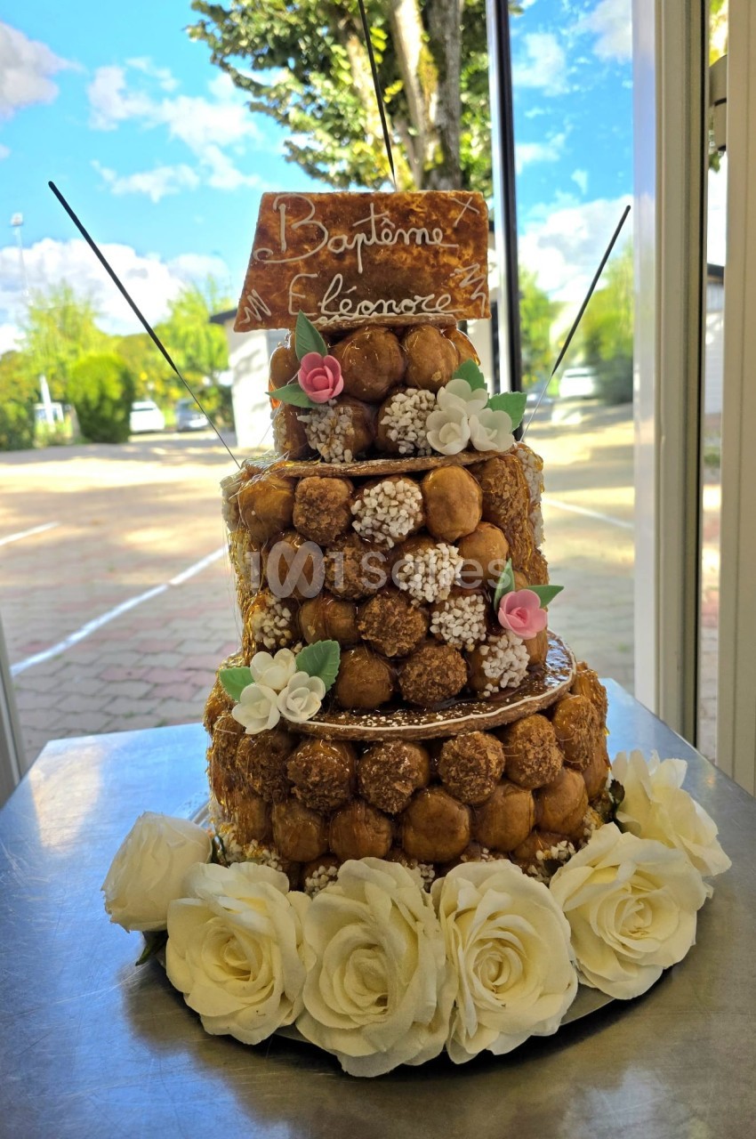 Pièce montée de choux décorée de roses blanches, fleurs en sucre et une plaque pour un baptême, posée sur une table.