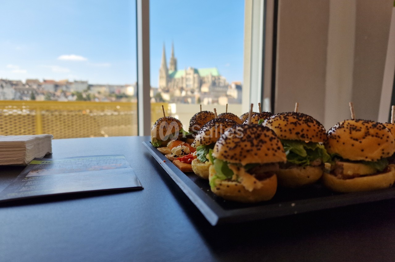 Plateau de mini-burgers garnis, posé sur une table près d'une fenêtre avec vue sur une ville et une cathédrale.
